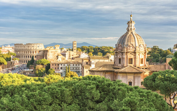 Panoramic view of Rome with the dome of Santissimo Nome di Maria al Foro Traiano and the Colosseum in the background, bathed in warm sunlight above lush green trees.
