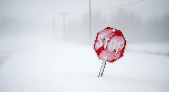 Stop sign covered in snow during blizzard conditions on empty road. Snowy landscape surrounds stop sign, highlighting winter weather effects.