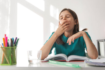 Tired schoolgirl studying at home yawns over an open book, showing fatigue overload from homework as sunlight falls across desk with stationery and notebooks creating a calm yet exhausted atmosphere