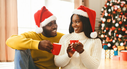 A couple sits together on the floor, wearing red hats. They smile and hold red cups filled with hot drinks. A decorated Christmas tree stands in the background.
