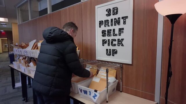 A man retrieves his completed 3D print project from a self-service pickup station inside a library workspace, with labeled bins and organized envelopes. 14975  