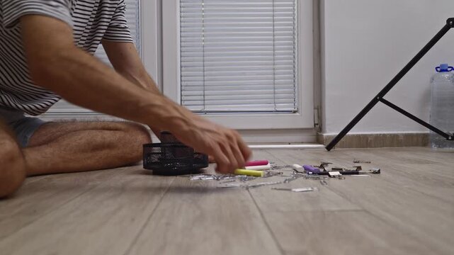 A close-up view of a person meticulously gathering scattered paper clips from a light wooden floor and placing them into a black mesh desk organizer.