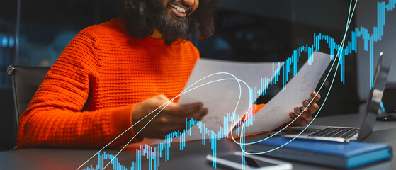 In a contemporary office setting, a cheerful businessman in a bright sweater examines important papers next to his laptop. His focus and smile reflect his satisfaction with the work.