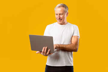 A person stands with a laptop in hands, looking at the screen. The background is bright yellow. The...