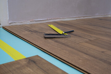 Laminate planks are being installed on a floor with a measuring square and tape. Precision is crucial for a smooth finish