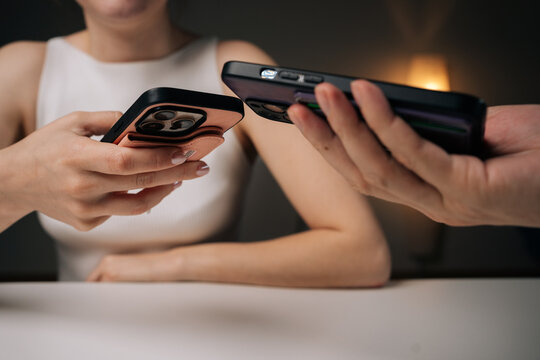 Two people exchanging data using mobile phones sitting at white table at home. Man and woman comparing smartphone data, sharing digital information in casual workspace environment.