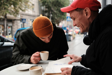 Two men review hand drawn sketches together at an outdoor table. The scene highlights creativity concept and collaboration concept.