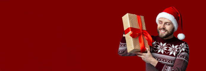 A cheerful man wearing a holiday sweater and Santa hat smiles as he holds a beautifully wrapped gift with a red ribbon. The vibrant red background adds to the festive spirit.