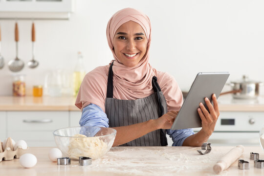 A woman wearing a pink hijab smiles as she holds a tablet in one hand in a cozy kitchen. Flour and baking tools surround her, indicating she is preparing to bake. - Powered by Adobe