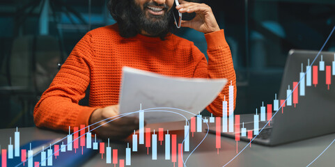 A cheerful young Indian man is seated at a table in a contemporary office space. He is on a phone call and reviewing marketing reports on his laptop, radiating confidence and success.