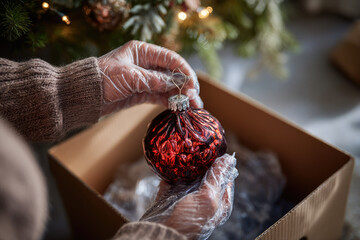 Taking down christmas tree decorations with care, hands wearing protective gloves gently placing red ornament into cardboard box, festive atmosphere, holiday season ends
