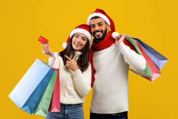 A joyful couple wearing Christmas hats smiles while holding colorful shopping bags and a red gift card. Their cheerful expressions reflect the festive holiday spirit and excitement of shopping.