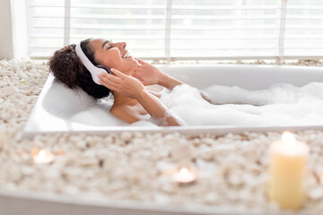 A woman enjoys a soothing bubble bath, wearing headphones and smiling. She relaxes in a stylish tub filled with bubbles and surrounded by candles, creating a tranquil atmosphere.