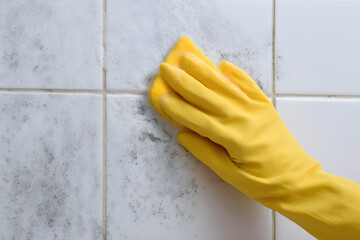 Woman in yellow glove scrubbing black mold from white tile. Household cleaning and bathroom maintenance concept for hygiene and fungus removal.