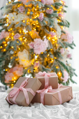 Beautifully wrapped Christmas present, gift boxes with soft pink ribbons lying on bed, with blurred festive lights and decorated tree in background, creating aesthetic, warm and elegant holiday mood.