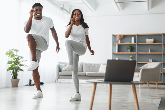 Online Family Workout. African American Husband And Wife Training Together In Living Room, Doing Twist Knees To Elbow Exercise. Happy Black Standing And Lifting Leg Up, Using Laptop, Watching Tutorial