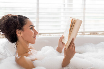 A person enjoys a relaxing moment in a bubble bath while reading a book. Natural light filters in through the blinds, creating a serene atmosphere.
