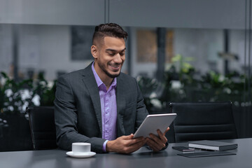 Fototapeta premium A man in a suit sits at a conference table looking at a tablet. He enjoys his work while surrounded by plant decor. A cup and notebook sit on the table.