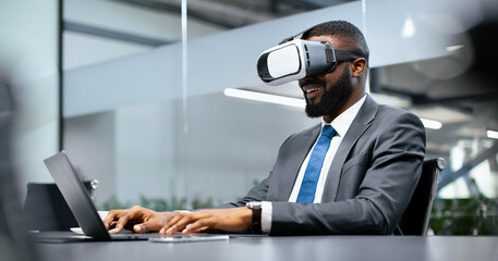 Businessman sits at a desk with a laptop open, wearing a virtual reality headset in a modern office. He smiles while interacting with the virtual environment during work hours.