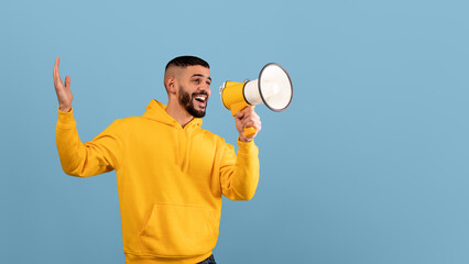 A man wearing a bright yellow hoodie is excitedly speaking into a megaphone. He is animated and cheerful, gesturing with his hands, set against a solid blue backdrop.