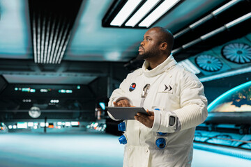 Orbit station crew member operating spacecraft control interface using tablet. Africana merican man wearing cosmonaut gear in spaceship doing research, monitoring system diagnostics