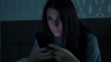 A young woman sits in a dark room at night, illuminated by her phones glow. She checks messages with deep focus, reflecting digital overload and emotional dependence. 