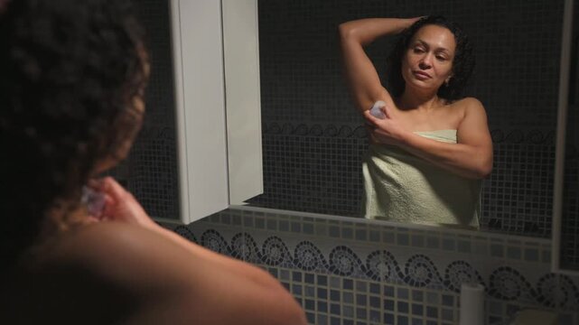 Young woman holing a natural crystal deodorant from alum salts, standing at bathroom sink, and looking at her reflection in the mirror. Eco-friendly and organic cosmetics for health and wellness