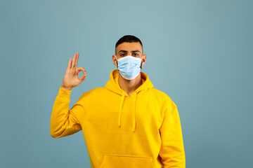 A young man wearing a bright yellow hoodie and a light blue mask stands against a light blue background. He is making an okay hand gesture, smiling confidently.