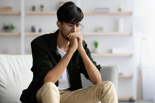 A young man sits on a gray couch, hands clasped together in front of his face. He appears contemplative, lost in thought in a well-lit living room with plants on shelves.