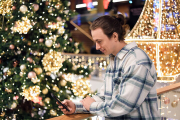  man using smartphone near Christmas tree in shopping mall. Happy male texting on phone against holiday lights background.