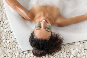 A woman enjoys a calming bubble bath while wearing a green mud mask. The serene atmosphere features white stones surrounding the tub, enhancing relaxation.