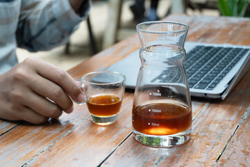 Close up of a glass beaker and shot glass containing dark coffee or tea on a rustic wooden table with a laptop.