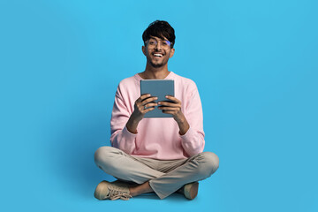A young man is sitting cross-legged on a bright blue background. He holds a tablet and smiles widely, enjoying what he is viewing on the device.