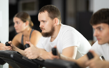 Obraz premium Bearded man hardly perform exercise on stationary bicycle, pedaling and exercising in fitness class. Fitness equipment, visitors engaged in physical task during training