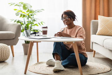 In a bright and warm living room, a young woman sits on the floor beside a coffee table, smiling...