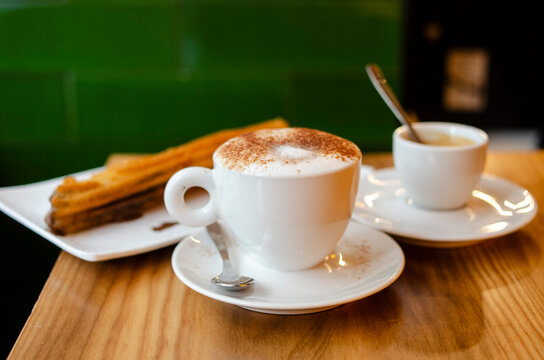 A trio of Spanish cafe treats -- a warm, frothy coffee with cinnamon, an espresso and a churro with chocolate on a wood table. Horizontal image.