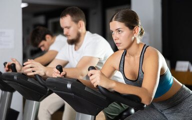 Obraz premium Woman in a sports top and leggings is exercising on an exercise bike in the gym. Visitor to a fitness club at a group cycling training tries and pedals