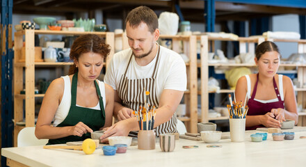 Fototapeta premium Two women craftsperson in striped aprons are busy molding clay products in lesson with male teacher. They are sitting near work table, sculpting plate of raw clay under supervision of teacher