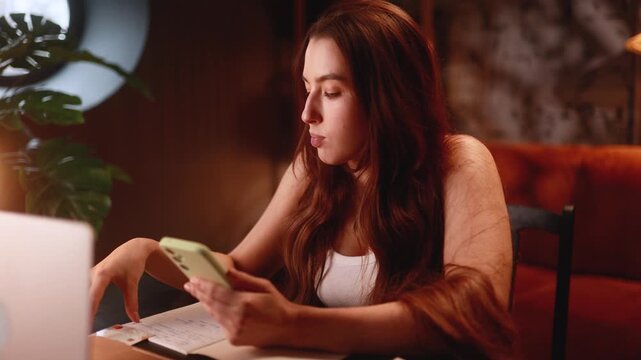 Beautiful woman at desk checking phone while studying late evening with laptop and notes, focused expression, warm lamp light, cozy apartment study scene showing concentration and quiet productivity