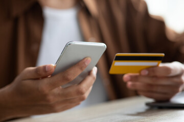 Individual is seated at a table, using a smartphone in one hand and a credit card in the other, about to complete an online transaction. Natural light shines through, creating a cozy atmosphere.