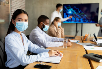 Diverse business professionals gather in a modern boardroom for a corporate meeting, practicing COVID-19 safety by wearing medical masks. Focus is on an Asian female employee engaged with the group.