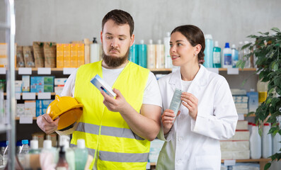 Obraz premium Male construction worker holding a package of cooling ointment for muscles and joints, he consults with a female pharmacist about choosing an ointment
