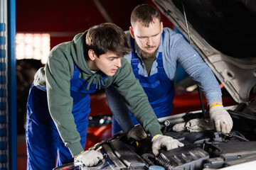 Fototapeta premium Two auto mechanics work together to diagnose an engine under the hood of a car in a repair shop