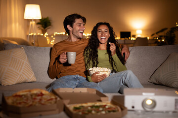 Cheerful couple relaxes on sofa, laughing while watching a comedy movie with a home cinema projector. They eat popcorn and pizza, enjoying their weekend indoors together.
