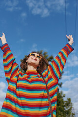 Woman in rainbow sweater wearing sunglasses, smiling with arms raised under blue sky. Young woman in her twenties, casual colorful knit and carefree pose in park for lifestyle fashion.