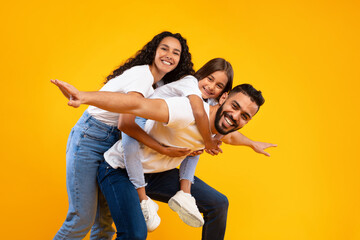 A happy family plays together in front of a vibrant yellow backdrop. A father carries his daughter on his back while the mother beams in delight, showcasing their bond and joy.