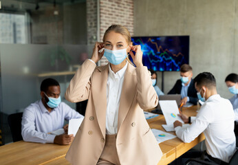 Young businesswoman adjusts her medical face mask before joining colleagues in the boardroom. Diverse team collaborates on projects during the coronavirus pandemic in a modern office setting.