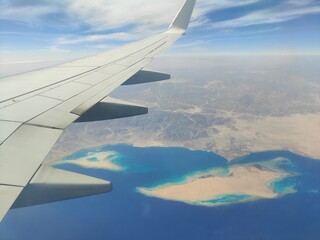 Obraz premium Aerial View of Airplane Wing Over Coastline and Sea, Red Sea, Egypt - A high-resolution aerial photograph taken from an airplane window, showing the aircraft wing above a deep blue sea with a desert c