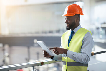 A person wearing a hard hat and safety vest is looking at a tablet while standing at a railing in a...