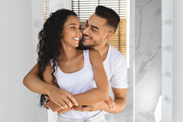 Obraz premium A young couple stands together in a stylish room, laughing and embracing each other. Sunlight filters through the slats, creating a warm atmosphere. Their happiness is evident.